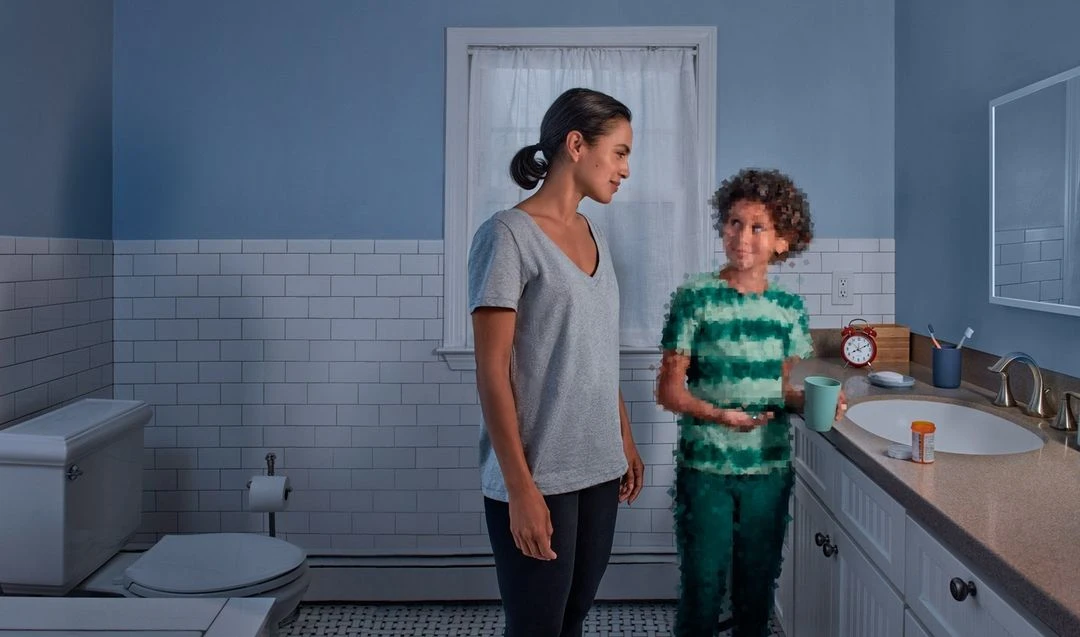 Mother and child in a blue-tiled bathroom with white trim. The child holds a cup and pill bottle near the sink.
